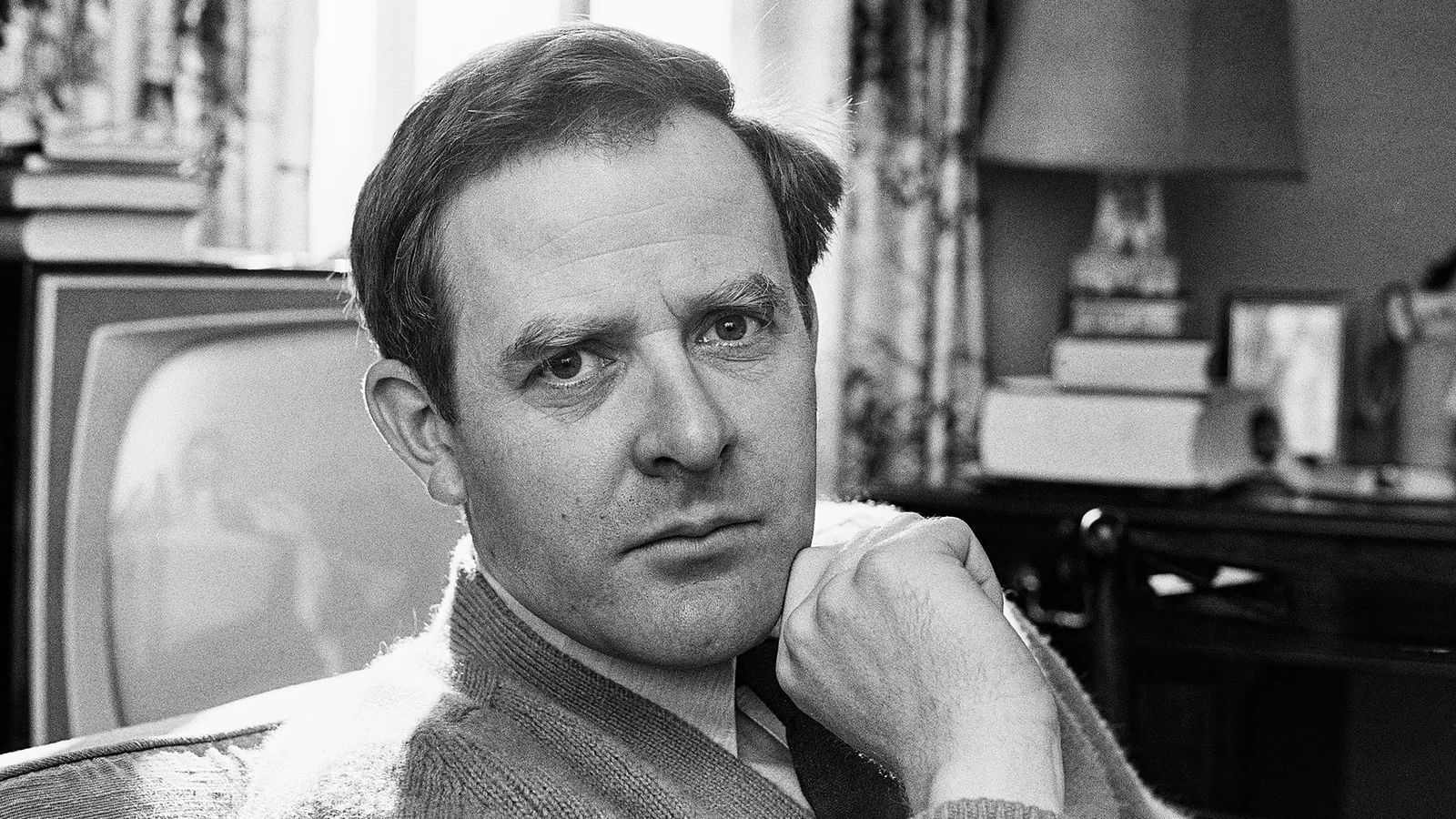 Black and white image of John le Carré sitting in a living room with a television behind him
